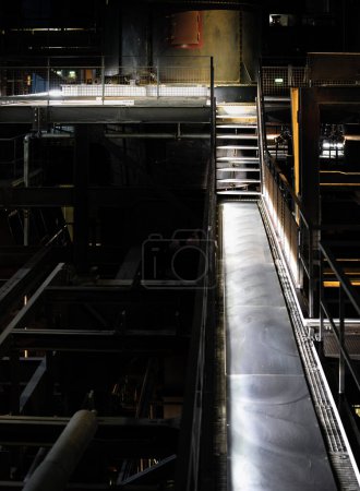 Un grand escalier métallique s'étendant vers le haut vers un bâtiment, donnant accès aux étages supérieurs. L'escalier est de conception industrielle, avec une construction simple et fonctionnelle.