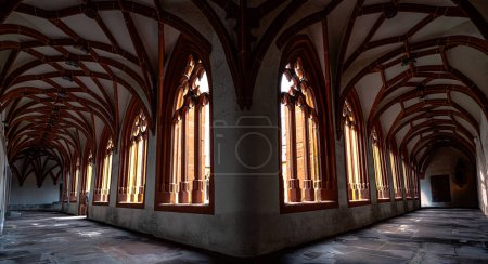 Die Sonne strömt durch die gewölbten Fenster und wirft einen warmen Schein auf den Steinboden dieses weitläufigen Korridors im gotischen Stil innerhalb eines alten Klosters..