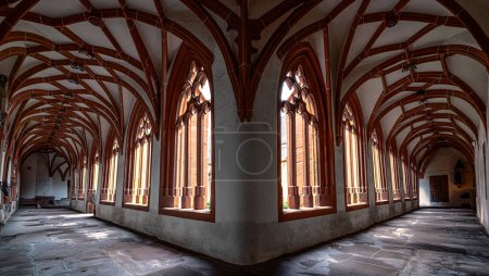 Le soleil coule à travers les fenêtres voûtées, projetant une lueur chaude sur le sol en pierre de ce vaste couloir de style gothique dans un ancien monastère.