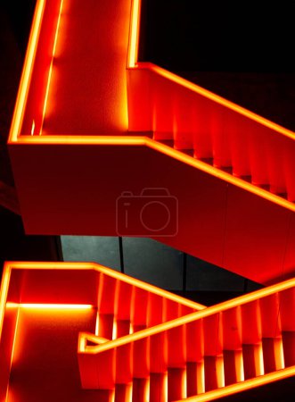 Lumière au néon rouge illuminant un ensemble d'escaliers, fournissant un guidage directionnel et une visibilité dans une zone sombre. Les escaliers mènent vers le haut, créant un sentiment de mouvement et de progression.