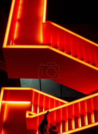 Luz de neón roja que ilumina un conjunto de escaleras, proporcionando orientación direccional y visibilidad en un área oscura. Las escaleras conducen hacia arriba, creando una sensación de movimiento y progresión.