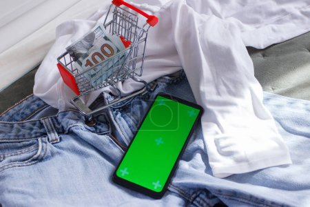 Foto de Carrito de compras con dólares americanos, un smartphone con pantalla verde, y ropa diferente. Concepto de viajar o ir de compras - Imagen libre de derechos