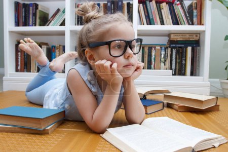 Foto de Little cute girl in the glasses sitting in the front of bookshelf. Concept of education in the library - Imagen libre de derechos