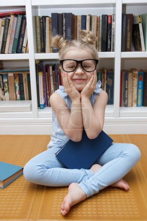 Foto de Niña linda en las gafas sentadas en la parte delantera de la estantería. Concepto de educación en la biblioteca - Imagen libre de derechos