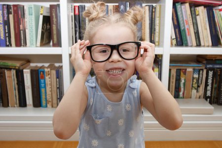 Téléchargez les photos : Petite fille mignonne dans les lunettes assis à l'avant de la bibliothèque. Concept d'éducation à la bibliothèque - en image libre de droit