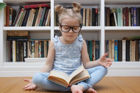 Téléchargez les photos : Little cute girl in the glasses sitting in the front of bookshelf. Concept of education in the library - en image libre de droit