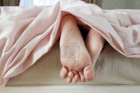 Téléchargez les photos : Pieds sous une couverture légère sur le lit, fond de mise au point doux. Concept de santé - en image libre de droit