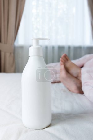 Photo for White bottle with a cosmetic product on the background of feet under a light blanket on the bed, soft focus background - Royalty Free Image