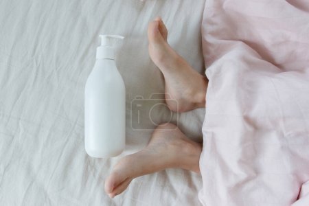 Téléchargez les photos : Flacon blanc avec un produit cosmétique près des pieds sous une couverture légère sur le lit, fond doux - en image libre de droit
