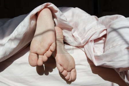 Füße unter einer hellen Decke auf dem Bett, weicher Hintergrund. Gesundheitskonzept