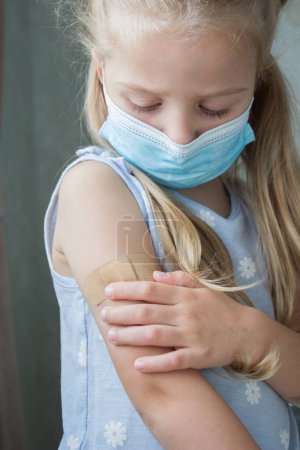 Téléchargez les photos : Petite fille dans un masque tient un bras avec un plâtre sur la peau. Concept de vaccination et d'immunisation - en image libre de droit