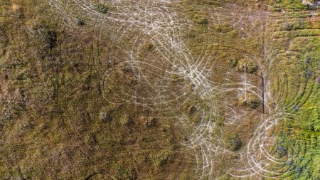 Vista aérea de un campo de fútbol abandonado. Un poste de gol solitario se encuentra en medio de la hierba demasiado grande. Pistas circulares de motos cruzan el campo, indicando donde la naturaleza y la actividad humana se cruzan.