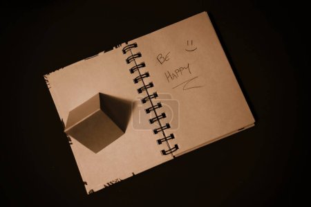 notepad with pages recycled from natural products, with the writing "be happy" and a smiling face, on the side a small pyramid made of the same material as the pages, which cast a shadow like an arrow indicating the writing