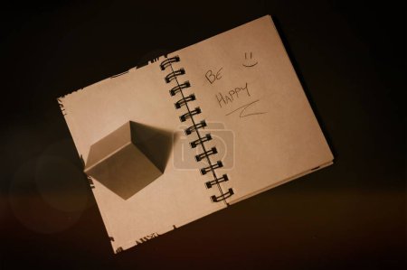 notepad with pages recycled from natural products, with the writing "be happy" and a smiling face, on the side a small pyramid made of the same material as the pages, which cast a shadow like an arrow indicating the writing