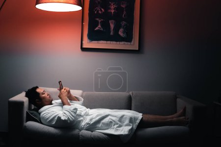 Schläfriger Mann, der im Bett liegt, abhängig von seinem Handy, Symptome von Schlafstörungen oder Rachegelüsten
