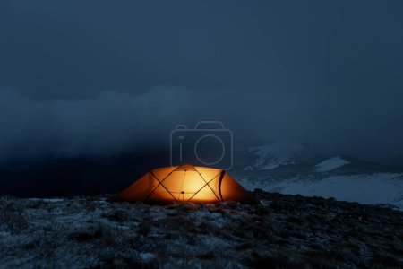 An orange tent glows warmly in a foggy mountain landscape during nighttime, creating a stark contrast with the chilly, desolate surroundings.
