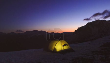 Ein leuchtendes Zelt steht auf einem schneebedeckten Berg unter einem dämmrigen Himmel, umgeben von schönen, heiteren Gipfeln in der Ferne.
