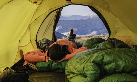 Une personne vêtue d'une veste orange repose dans une tente jaune, regardant les montagnes lointaines par temps clair.