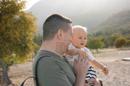 Photo for Dad holds his daughter in his arms, hugs and talks to her, beach and beautiful background with trees and mountains at sunset. High quality photo - Royalty Free Image