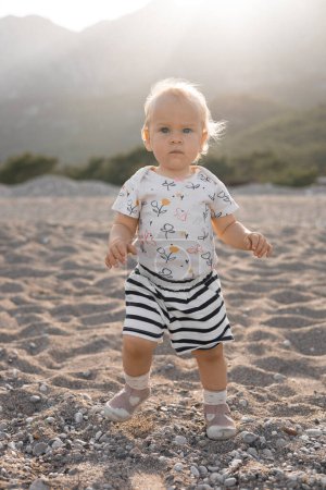 Photo for Beautiful little blonde girl in shorts and a T-shirt plays on the beach, happy sunny spring day. High quality photo - Royalty Free Image