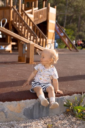 a little girl in shorts and a T-shirt sits on the floor against the background of a childrens playground, hot spring. High quality photo