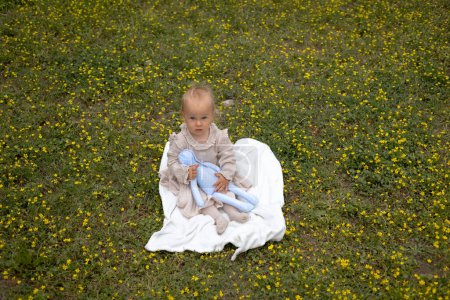 Photo for Cute little girl in a beige dress sits on a blanket and plays with a soft hare in yellow flowers. - Royalty Free Image