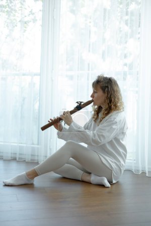 Photo for Beautiful young curly woman playing a wooden flute at home near the window, wearing in white outfit - Royalty Free Image