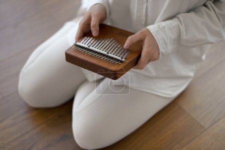 Kalimba in den Händen einer jungen Musikerin in weißen Kleidern, die zu Hause auf dem Boden sitzt