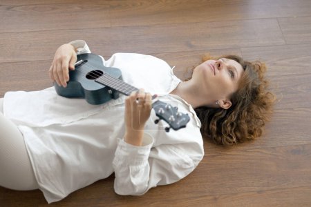 Photo for Young beautiful curly woman plays the ukulele, lying on the floor in a bright studio in white clothe - Royalty Free Image