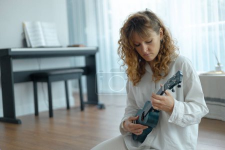 Photo for Portrait of a young beautiful curly woman plays the ukulele, she sits on the floor in a bright studio in white clothe - Royalty Free Image