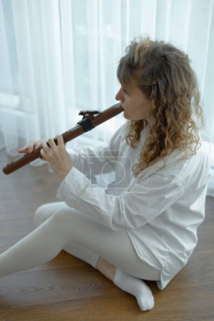 Photo for Beautiful young curly woman playing a wooden flute at home near the window, wearing in white outfit - Royalty Free Image