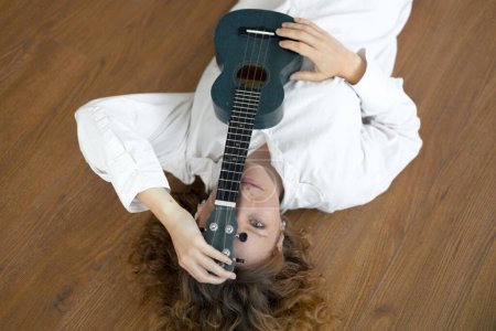 Photo for Portrait of a young beautiful curly woman with ukulele covered a half of her face, she lyimg on the floor in a bright studio in white clothe - Royalty Free Image