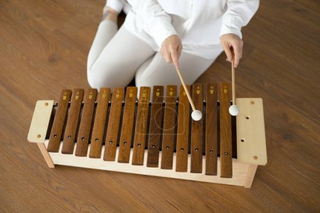 Beautiful young curly woman playing on a wooden xylophone sitting on the floor in white clothes.
