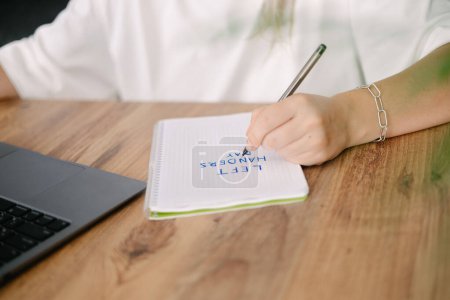 Frau schreibt mit der linken Hand in ein Notizbuch den Text, den Linkshänder Tag. Holztisch, grüne Blume und Laptop auf Hintergrund
