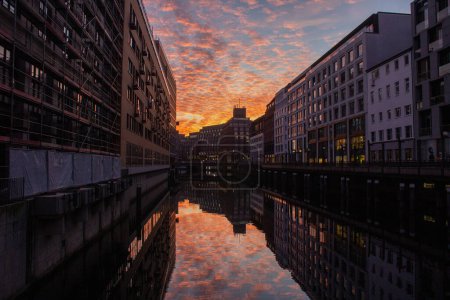 A serene sunset reflects off the calm waters of an urban canal, flanked by modern buildings. The fiery sky mingles with soft clouds, casting a warm glow that contrasts with the cool blues of the cityscape.