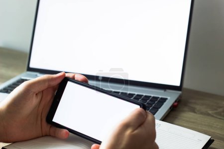 man hands with mobile phone and laptop on background, with blank white screens. business concept. mock up,blank white screens of mobile phone and laptop, man holding phone in front of laptop at the table, scrolling , notebook and pen nearby, close up
