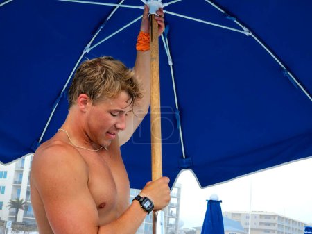 Millennial handsome guy working on beach setting up beach umbrellas. 