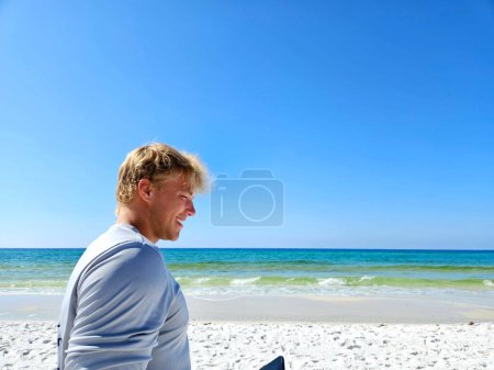 Young man on vacation walking down sandy beach using cellphone. 