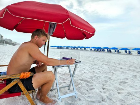 Millennial-Geschäftsmann arbeitet in seinem Büro unter rotem Regenschirm am Strand und verkauft Liegestühle und Sonnenschirme. 