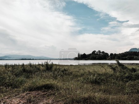 A dark and beautiful Scenic view of the mountain with green grass and a calm lake with cloudy blue sky background at tasik timah tasoh, perlis, malaysia.