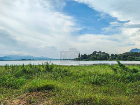 A beautiful Scenic view of the mountain with green grass and a calm lake with cloudy blue sky background at tasik timah tasoh, perlis, malaysia.