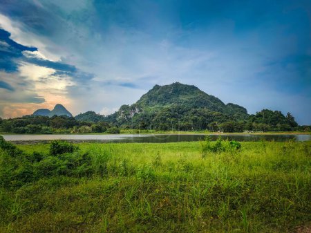 A dark and beautiful Scenic view of the mountain with green grass and a calm lake with cloudy blue sky background at tasik timah tasoh, perlis, malaysia.