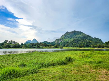 A beautiful Scenic view of the mountain with green grass and a calm lake with cloudy blue sky background at tasik timah tasoh, perlis, malaysia.