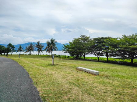 Schöne Aussicht auf den Park mit Bäumen, grünem Gras, einem See in Führzügeltechnik und einem wolkenverhangenen Himmel bei Tasik timah tasoh, Peru, Malaysia.