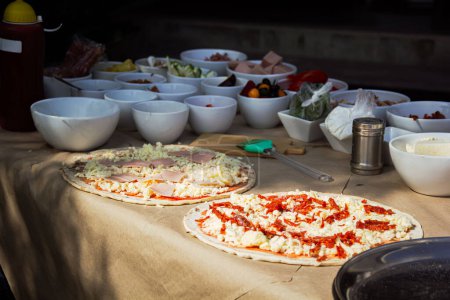 Téléchargez les photos : Une table remplie d'ingrédients - origan, huile d'olive, sauce chili, sauce tomate et pâte à pizza pepperoni - prépare le terrain pour un délicieux pique-nique à pizza dans le parc. Baignée de soleil - en image libre de droit