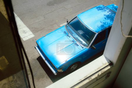 Captured from a window on a bright sunny day, this photo showcases a striking blue car illuminated by the warm rays of the sun, adding a vibrant touch to the scene