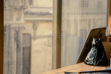 Un compagnon félin en céramique, tenant une cigarette dans sa main, repose gracieusement à côté d'une fenêtre ensoleillée, ajoutant du charme à l'atmosphère artistique du studio de peinture