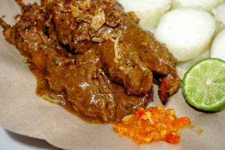 Nahaufnahme von Sate Ayam (Chicken Satay) mit Erdnusssoße und Lontong (Reiskuchen). Eines der beliebtesten Streetfood Indonesiens. Serviert in weißem Teller auf weißem Tisch. Selektiver Fokus.