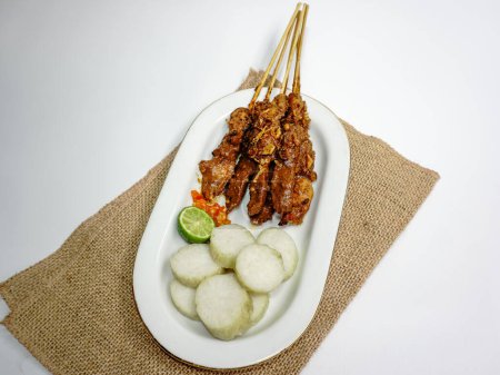 Sate Ayam (Chicken satay) with peanut sauce and lontong (rice cake). One of the popular street food in Indonesia. Served in white plate on white table. Selective focus.