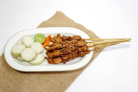 Sate Ayam (Chicken satay) with peanut sauce and lontong (rice cake). One of the popular street food in Indonesia. Served in white plate on white table. Selective focus.